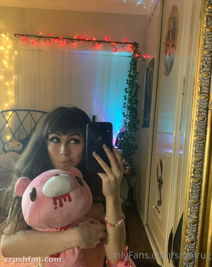 La créatrice aux longs cheveux foncés tient une peluche rose et un téléphone dans un selfie miroir, avec des guirlandes lumineuses rouges derrière.