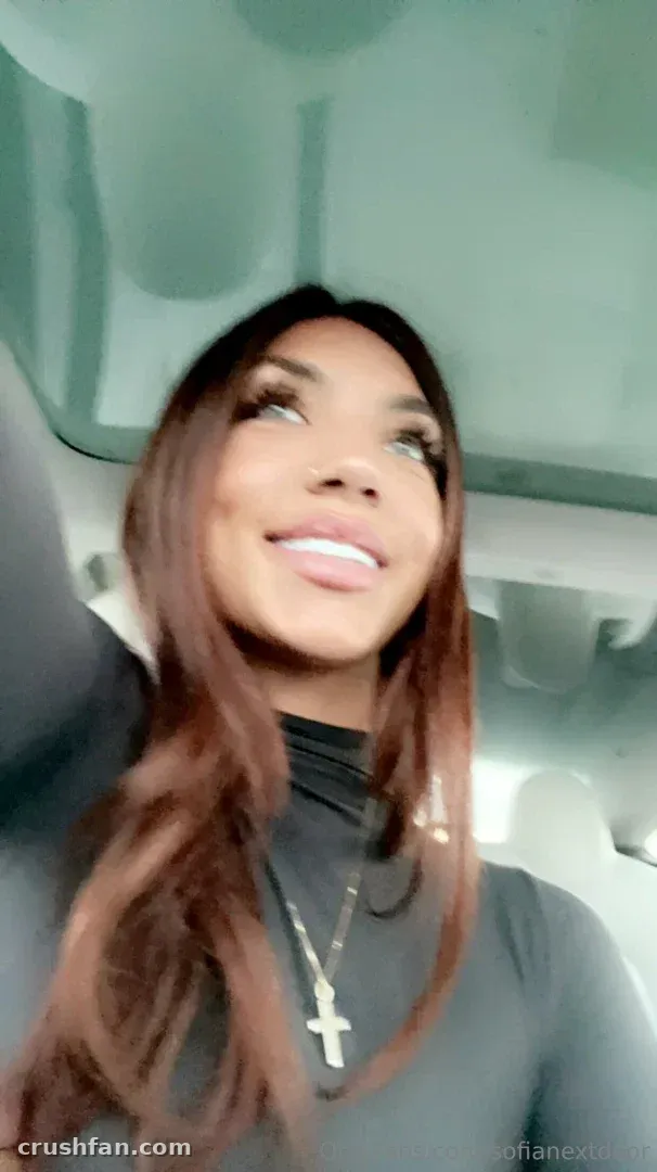 Sofianextdoor lächelt in einem Auto-Selfie, trägt ein dunkles Oberteil und eine goldene Kreuzkette.