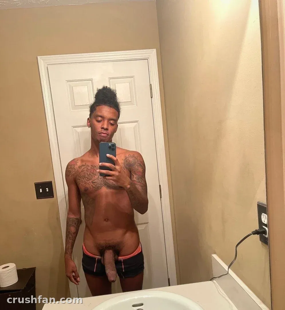 Shirtless tattooed man in dark shorts taking a mirror selfie in a beige bathroom.