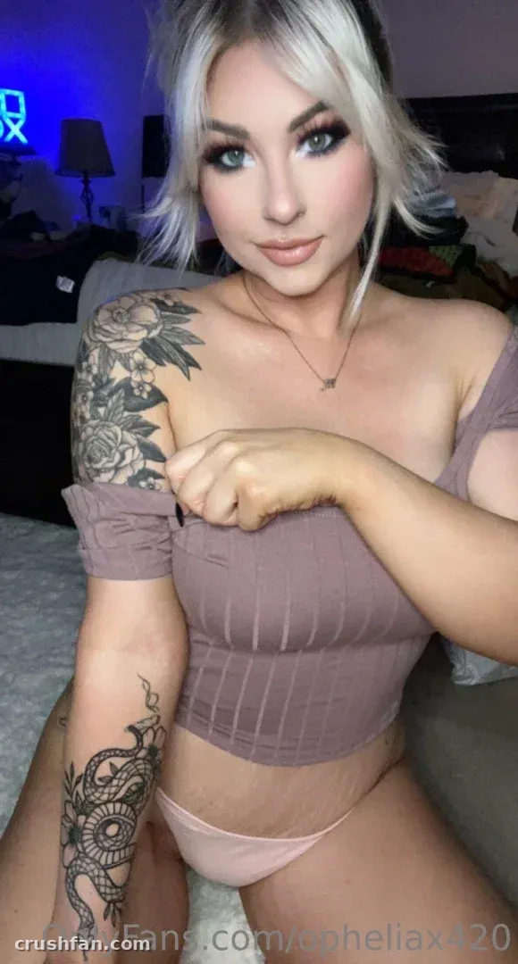 Ophelia “O” con un top malva de hombros descubiertos y tatuajes florales en el brazo en una habitación interior con poca luz
