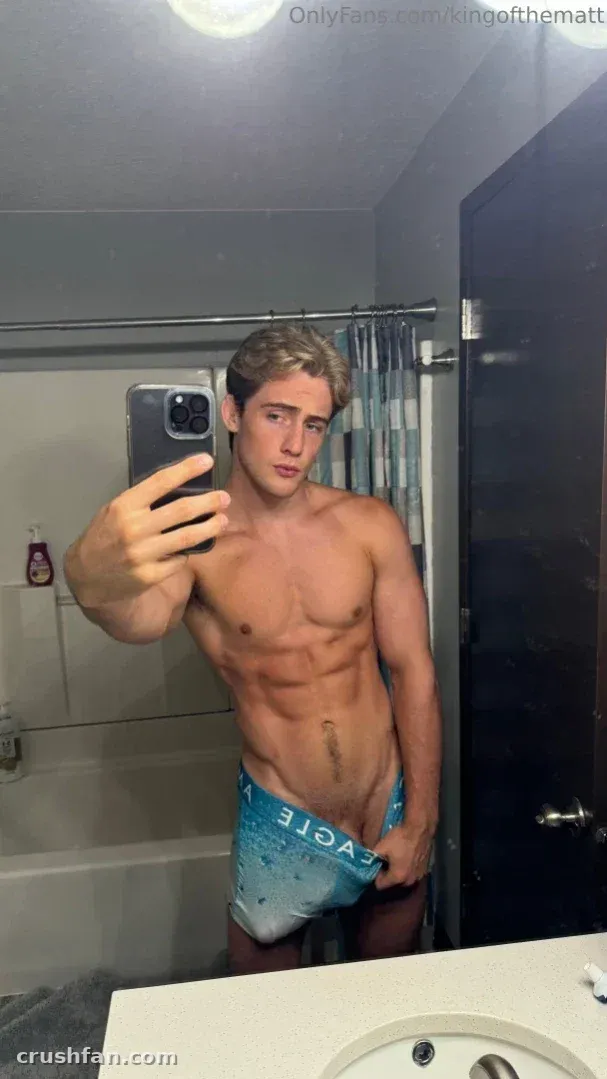 Homem sem camisa de sunga azul tirando uma selfie no espelho em um banheiro