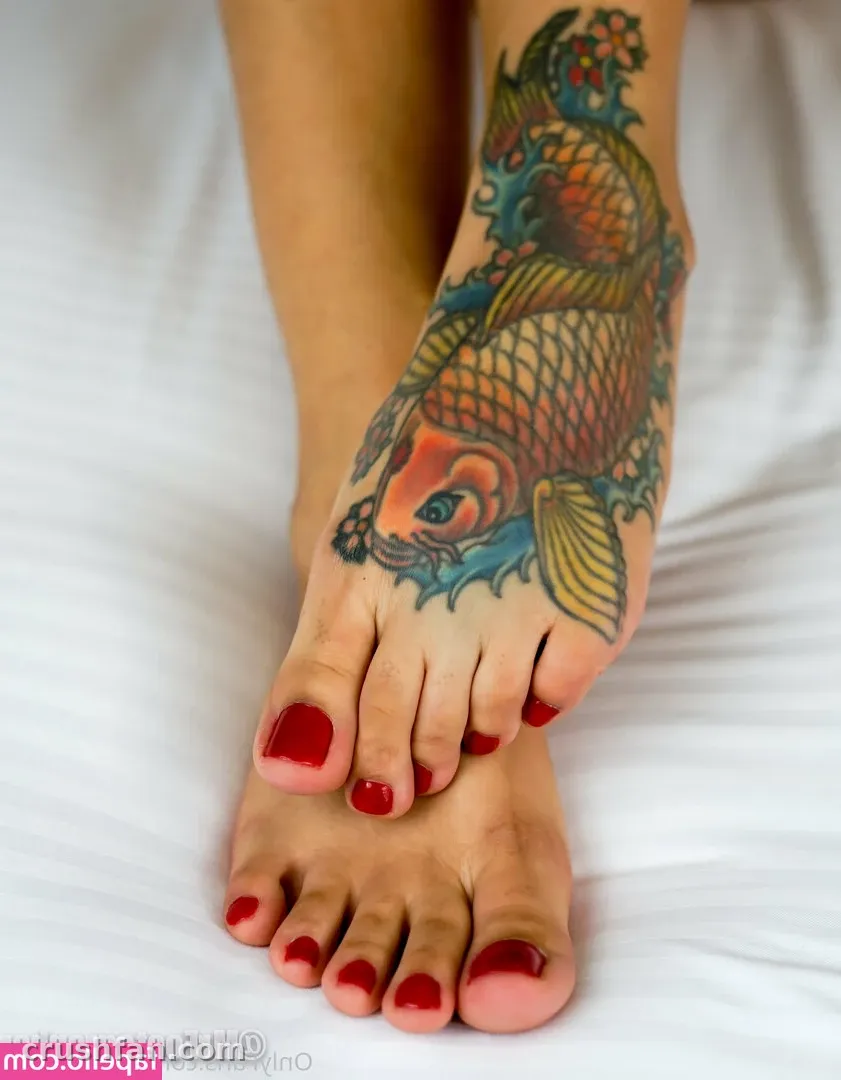 Close-up of a foot with a colourful koi fish tattoo and red toenails resting on white sheets