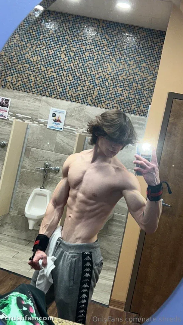 Nathan taking a mirror selfie in a tiled washroom wearing grey sweatpants and wrist wraps.