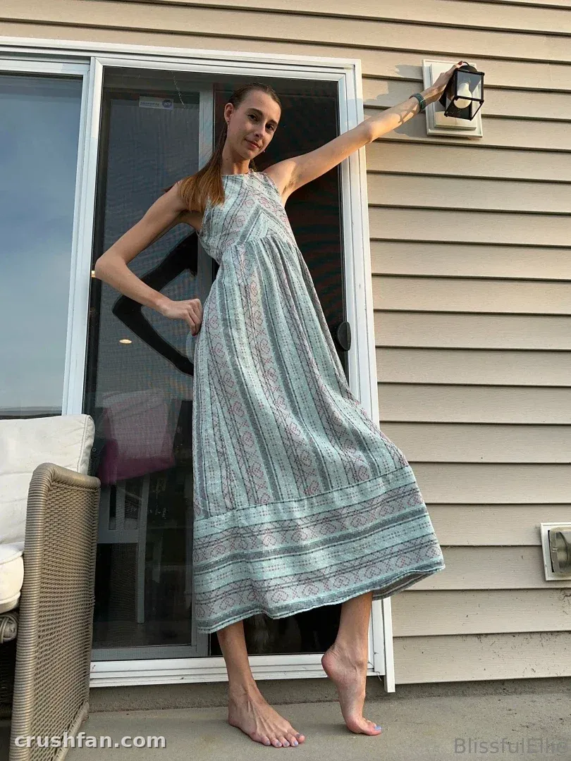 Ellie em um vestido longo listrado, descalça em um pátio perto de uma porta de vidro