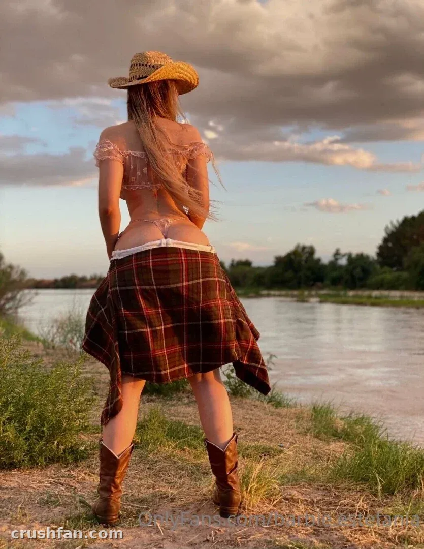 Mujer con sombrero de paja, blusa con volantes, falda de cuadros y botas vaqueras de pie junto a un río al atardecer