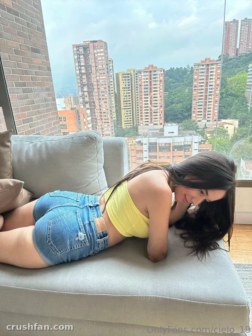 Mulher de top amarelo e shorts jeans deitada em um sofá cinza perto de uma janela