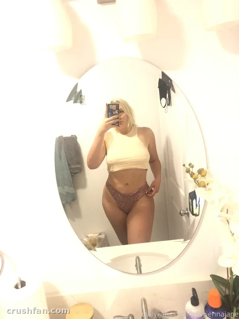 Jenna Jane prenant un selfie miroir dans une salle de bain lumineuse, portant un haut crème et une culotte de bikini à motifs.