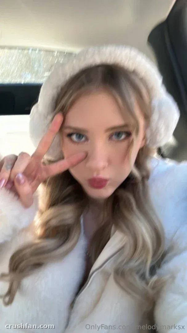 Melody Marks mit einem weißen Baskenhut und Pelzmantel, die im Inneren eines Autos ein Peace-Zeichen macht.