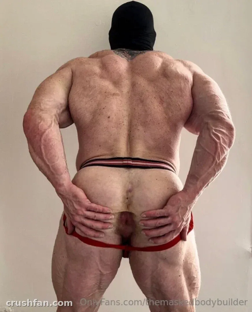 Muscular masked man seen from behind, hands on hips, against a plain wall