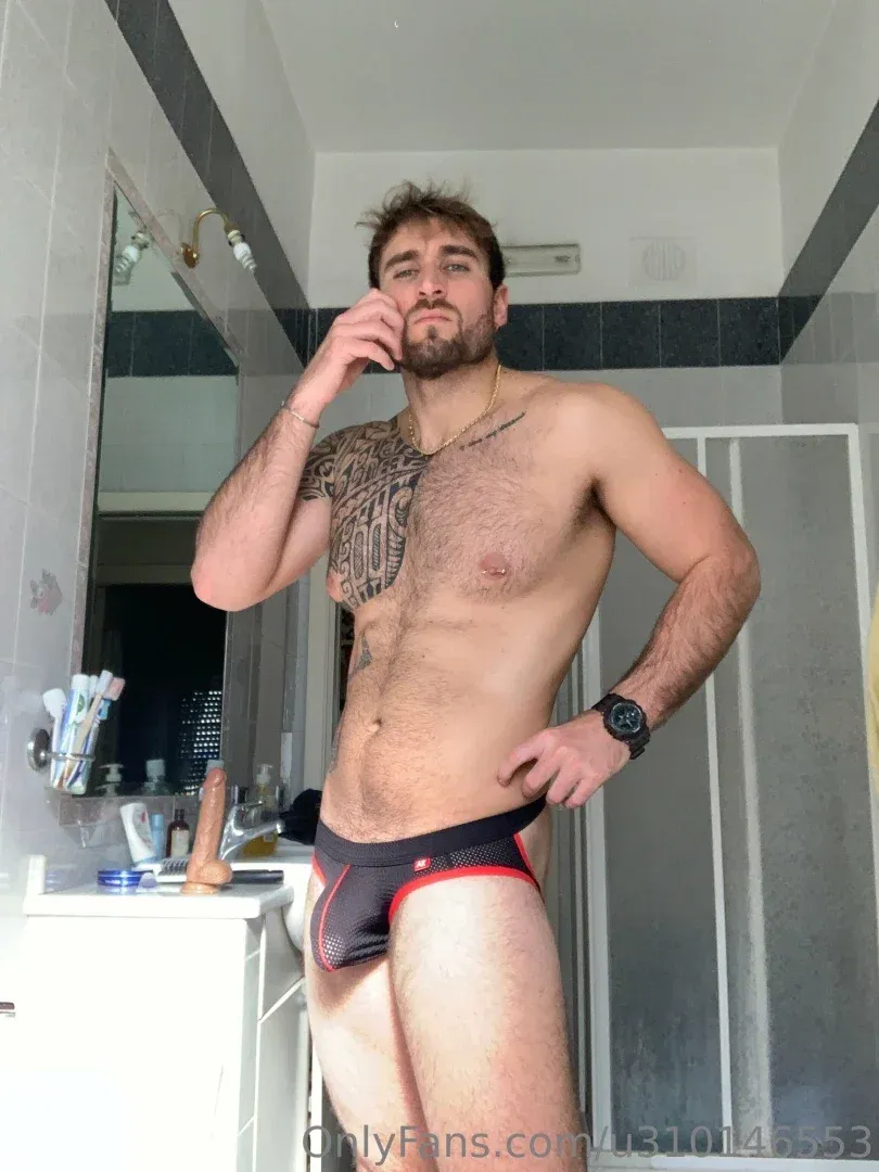 Shirtless man with chest tattoo taking a mirror selfie in a bathroom.
