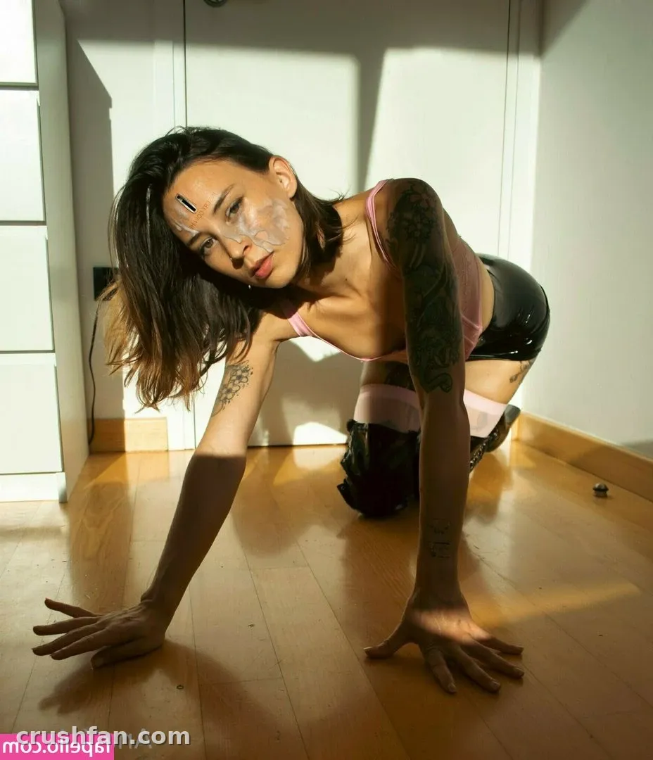 AnaKatana agachada em um piso de madeira com um top rosa e shorts pretos, com sombras dramáticas atrás dela.