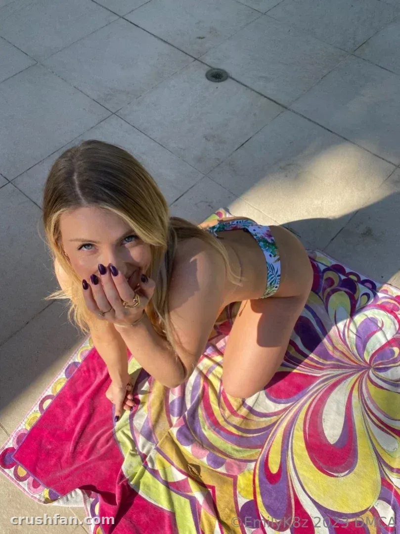 Emma K crouching on a colourful towel on grey paving stones in bright sunlight