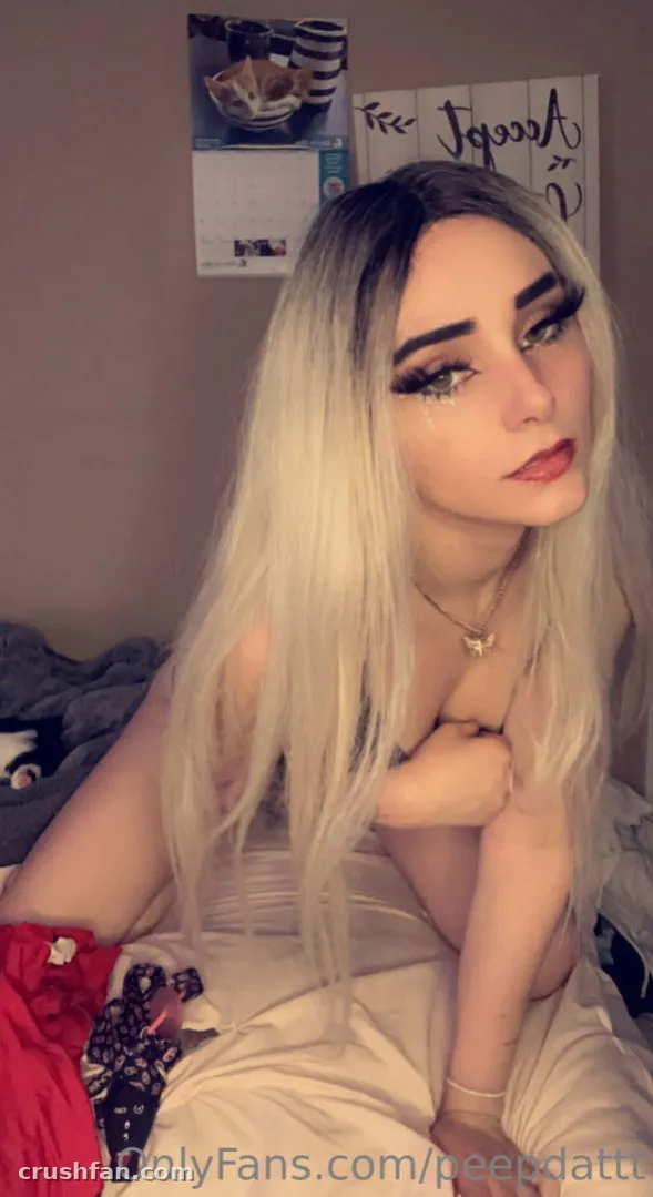 Creator aux longs cheveux blonds et au maquillage des yeux audacieux, assise en intérieur contre un mur avec une affiche.