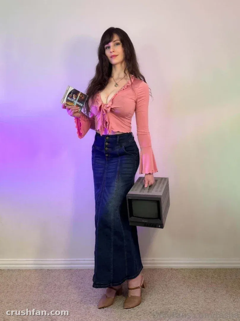 Tasha com um top rosa e saia jeans segurando uma caixa e uma televisão retrô