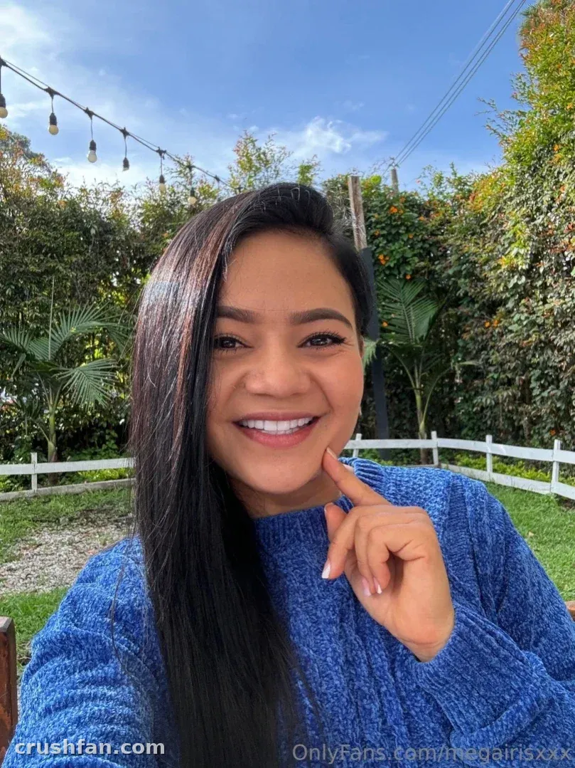 Megan Iris lächelt im Freien in einem blauen Strickpullover mit Grünpflanzen hinter ihr
