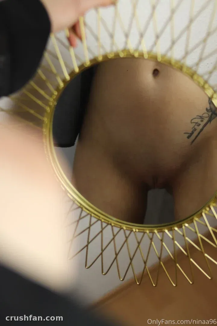 Nina Joy refletida em um espelho redondo de arame dourado, mostrando uma cintura tatuada e uma manga preta.
