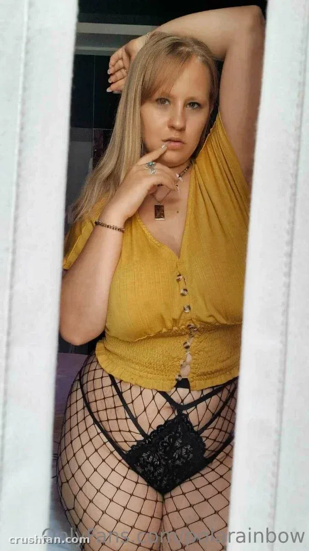 Creator in a mustard top and fishnet tights standing between pale curtains indoors