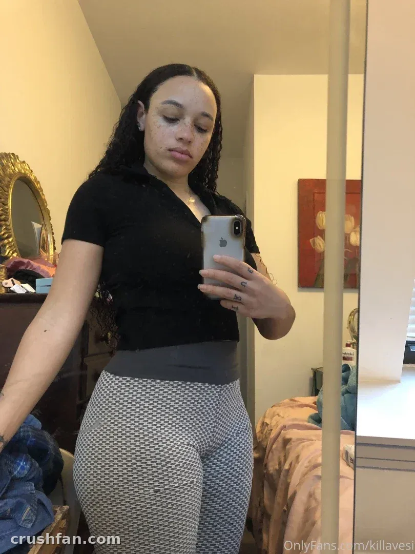 Killavesi in a black top and patterned leggings taking a mirror selfie in a bedroom