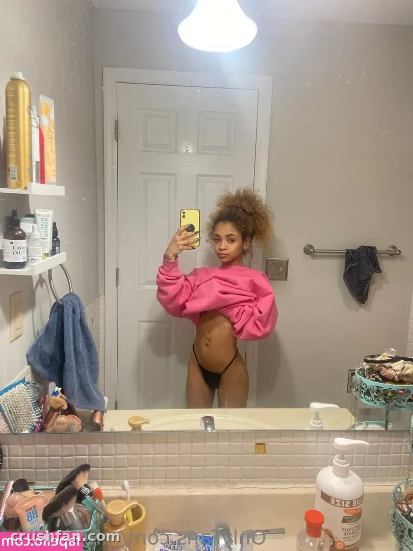 KITTY en sweat court rose et culotte de bikini foncée prenant un selfie dans le miroir dans une salle de bain.