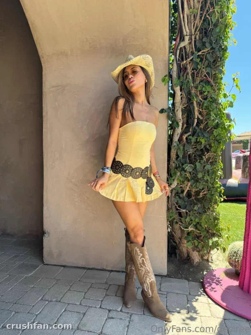 Criadora com vestido tomara que caia amarelo e chapéu de palha em pé junto a uma parede iluminada pelo sol com trepadeiras