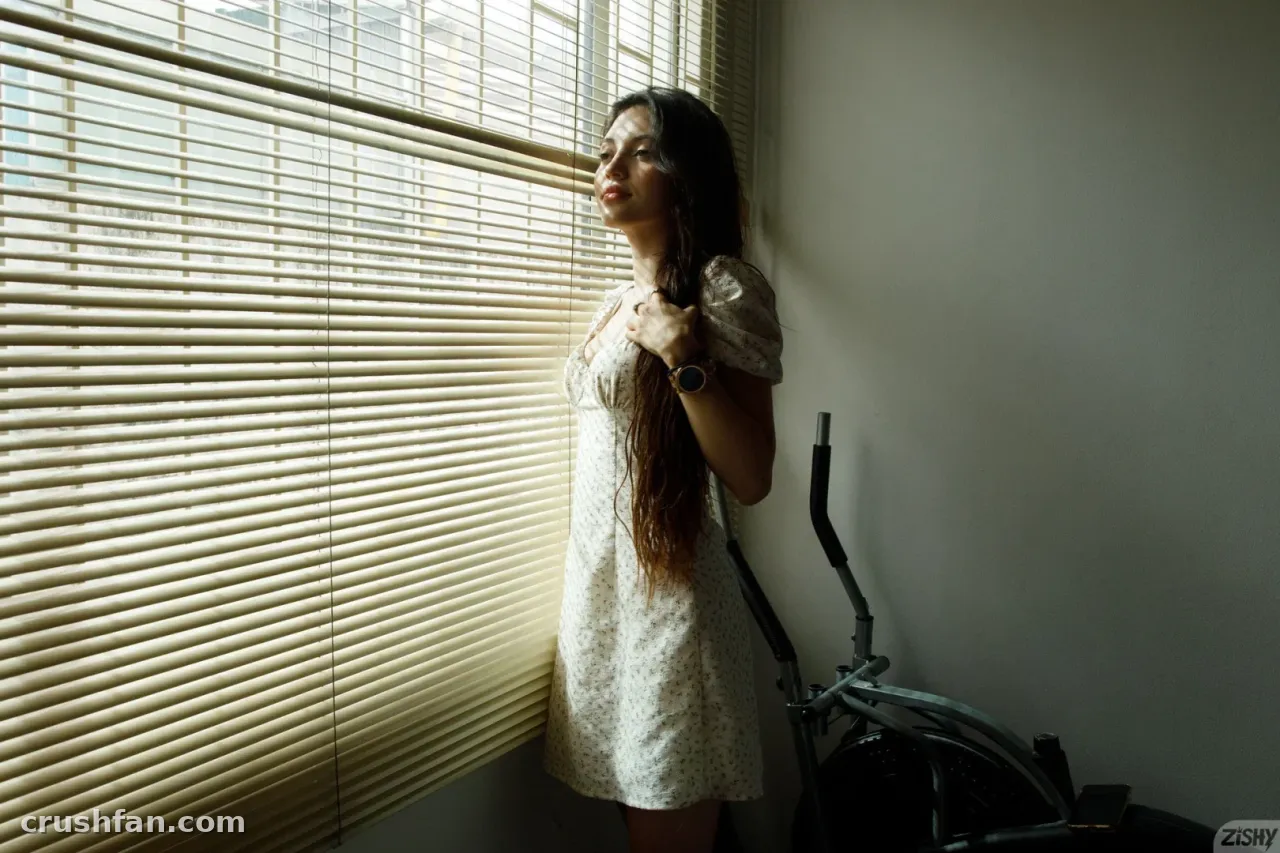 Ange in a white dress standing by closed blinds with warm light on her face
