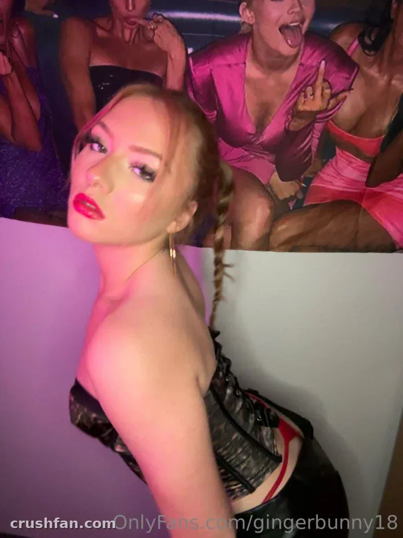 Creator GingerBunny in a strapless top posing in front of a pink-lit printed backdrop.