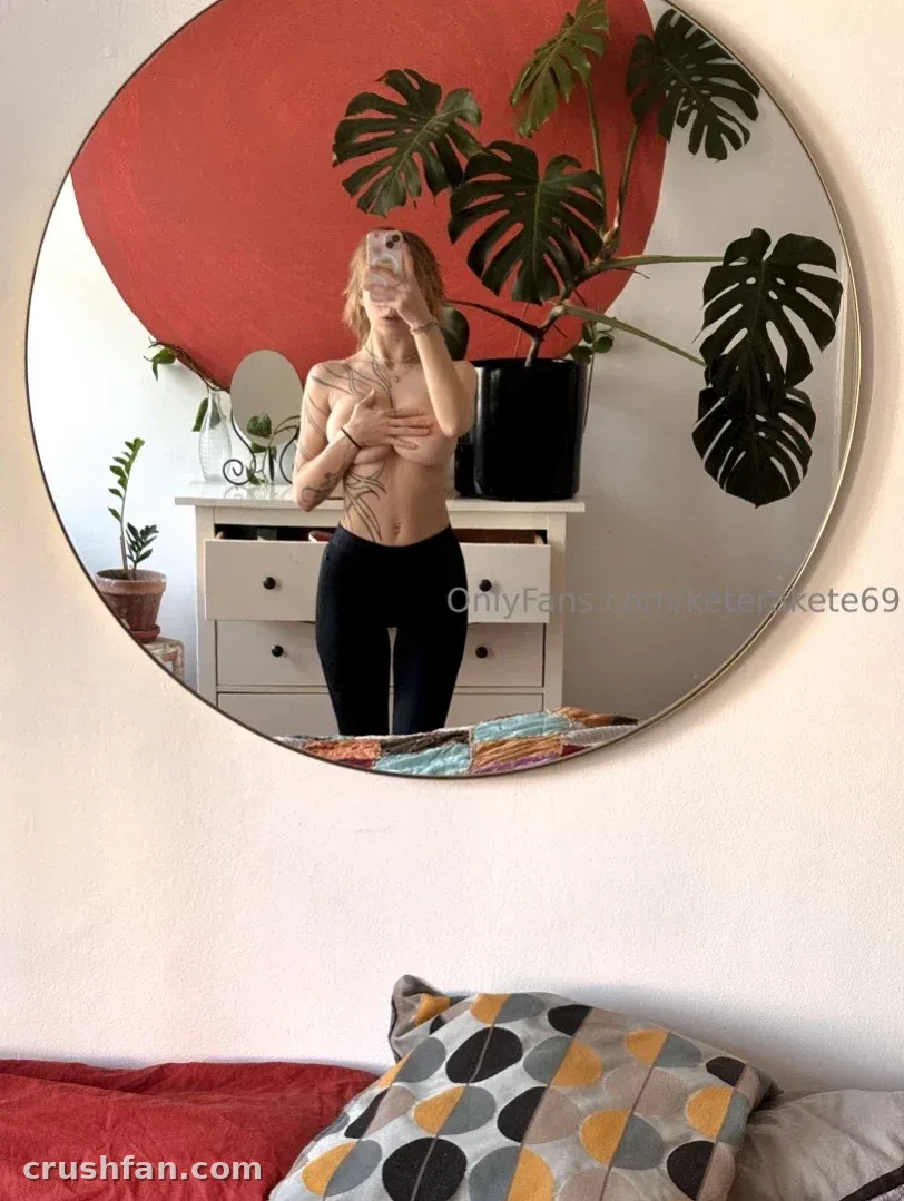Kat tirando uma selfie no espelho em um quarto com uma planta monstera e um painel de parede vermelho