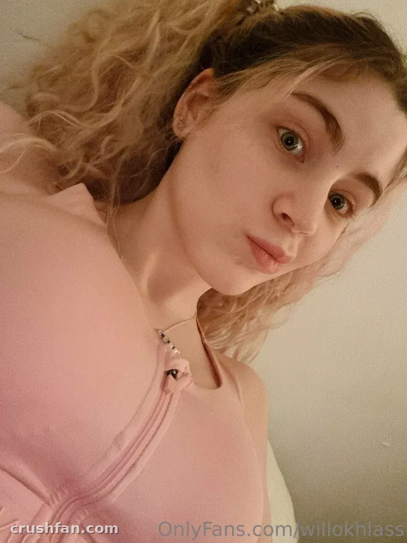 Willokhlass con una blusa rosa tomándose una selfie en primer plano en interiores contra una pared lisa