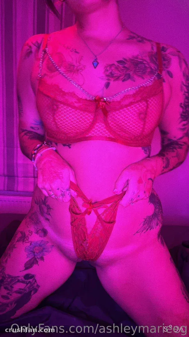 AshleyMarie en lingerie en filet rouge sous un éclairage rose, montrant des tatouages et un collier pendentif.