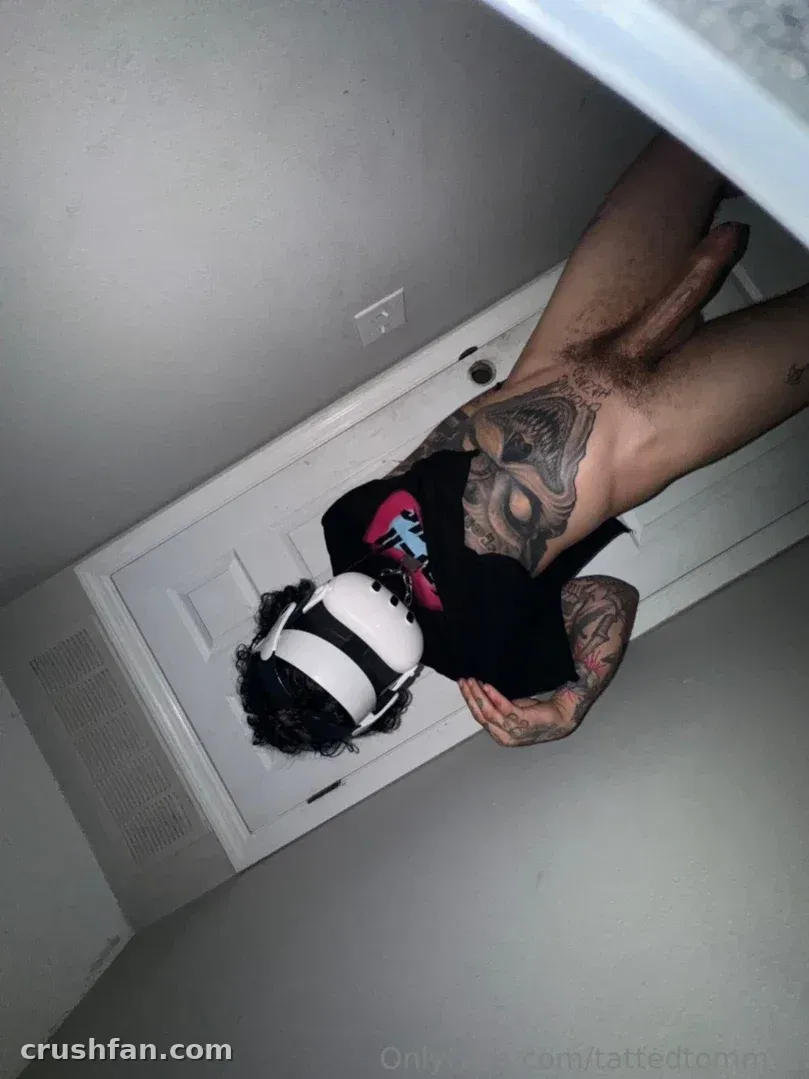 Tommy deitado em um piso cinza com uma camiseta gráfica preta e capacete branco em ambiente interno.