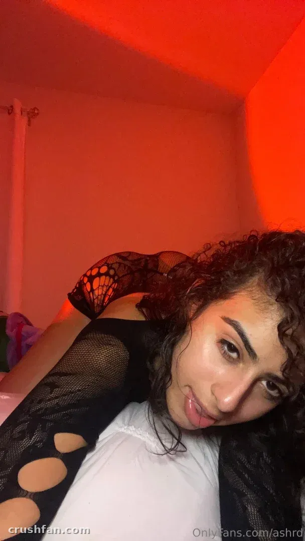 Ash in a black lace top looking at the camera under red lighting indoors