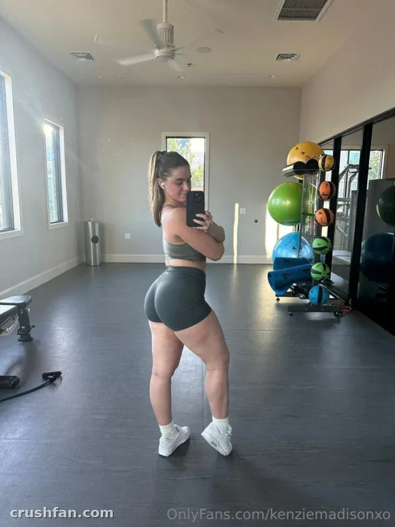 Kenzie Madison in grey activewear taking a mirror selfie in a bright home gym with exercise balls.