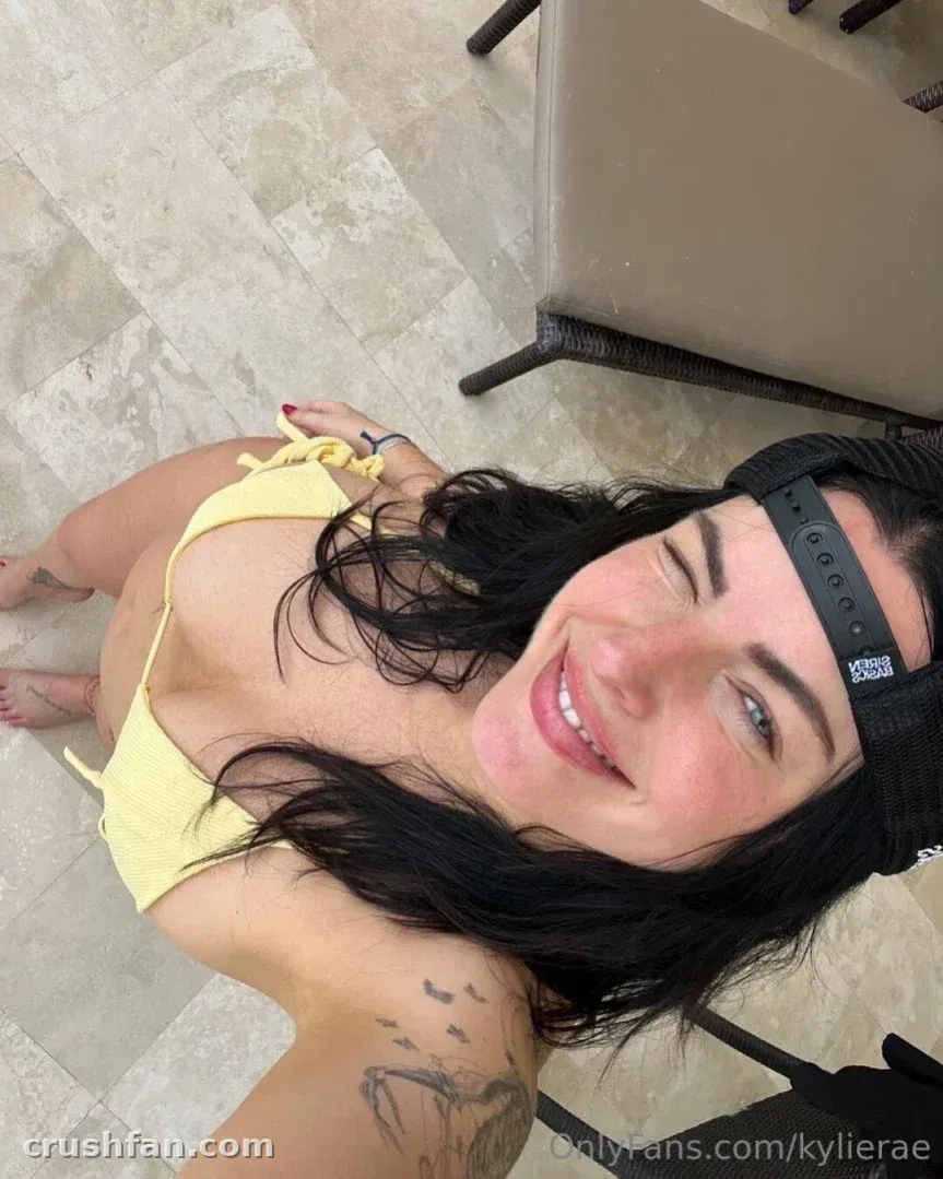 Kylie Rae con un top de bikini amarillo y una gorra hacia atrás, sonriendo hacia la cámara en un suelo de piedra.