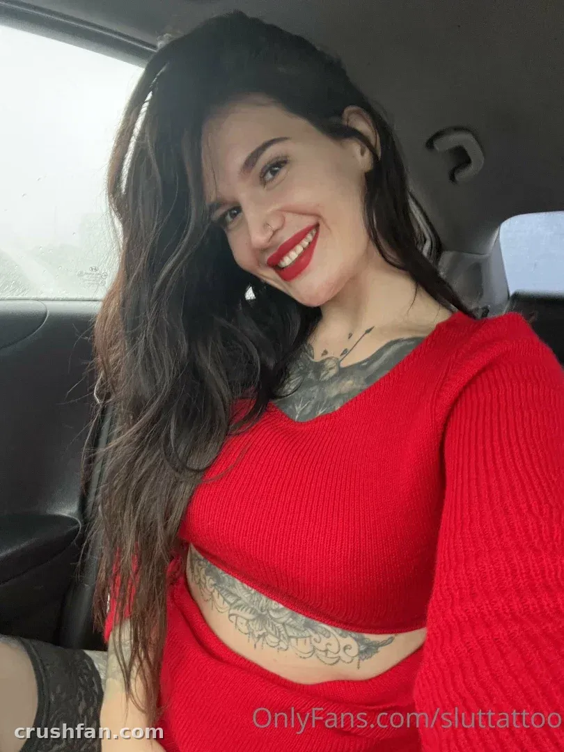 Smiling woman in a red ribbed crop top seated inside a car, showing chest tattoos.
