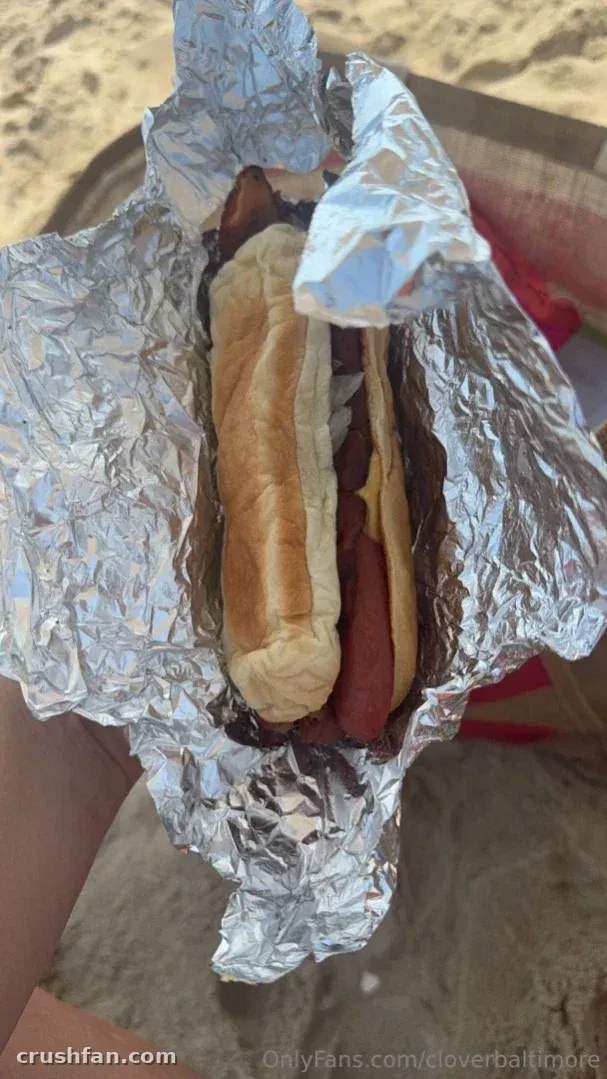 Cloverbaltimore holding a foil-wrapped hot dog with mustard at the beach