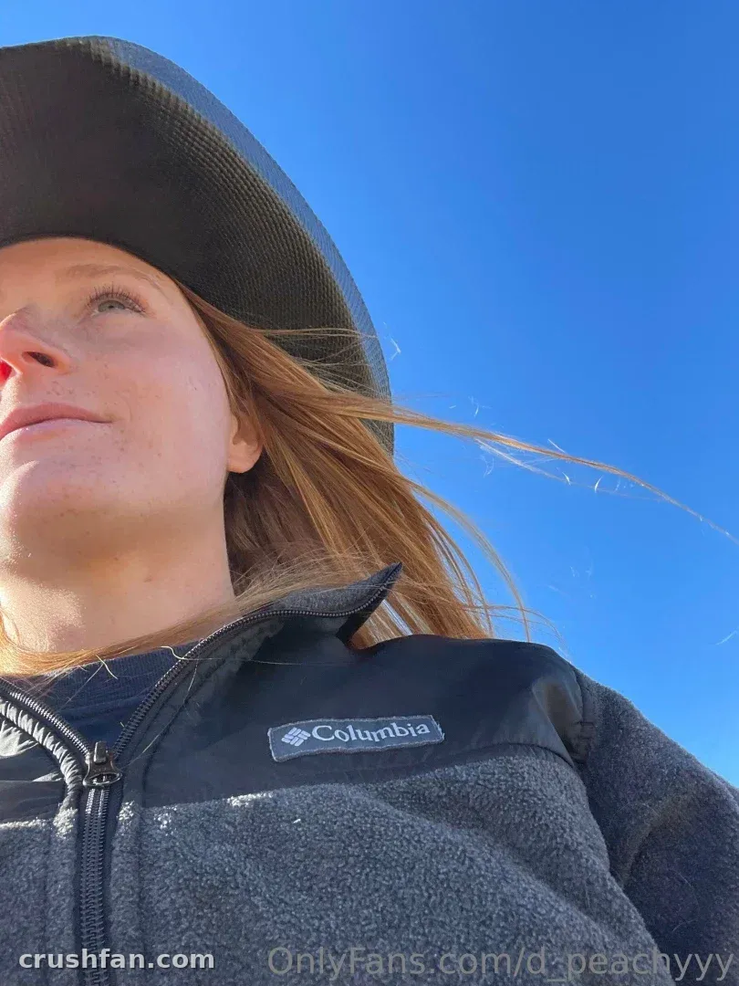 Low-angle outdoor portrait of a woman in a wide-brim hat and grey fleece against a clear blue sky