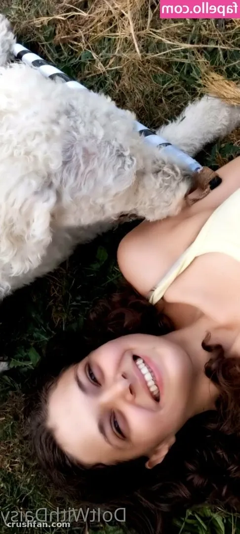 Smiling woman with long dark wavy hair lying on grass next to a fluffy white dog