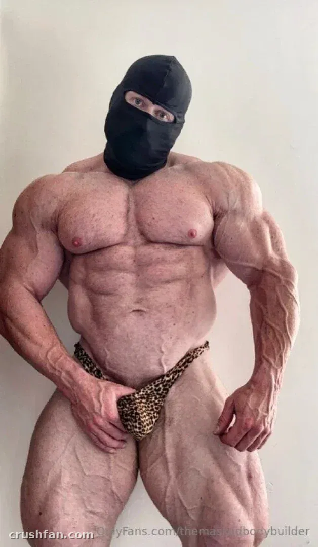 Muscular masked creator posing indoors in a leopard-print pouch against a plain wall.