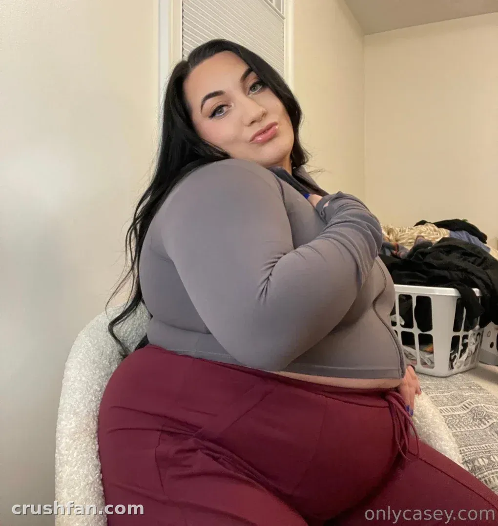 Casey com uma blusa cinza e calça vinho sentada em ambiente interno perto de um cesto de roupa