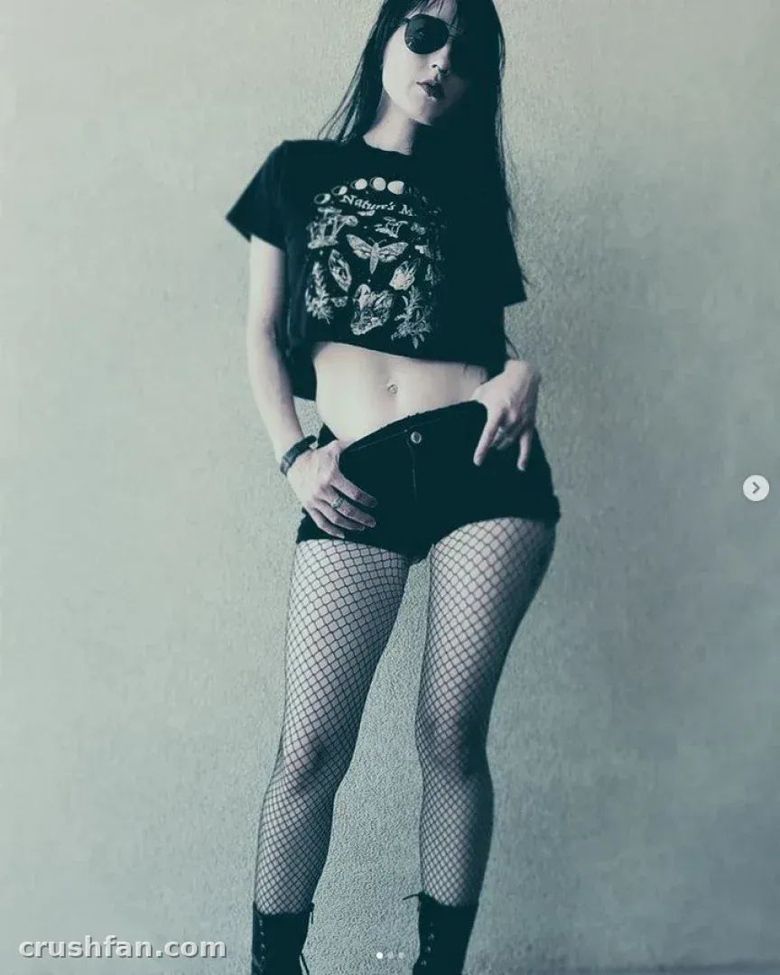 Freya in einem bauchfreien Grafik-T-Shirt, dunkler Shorts und Netzstrumpfhose vor einer schlichten Wand.
