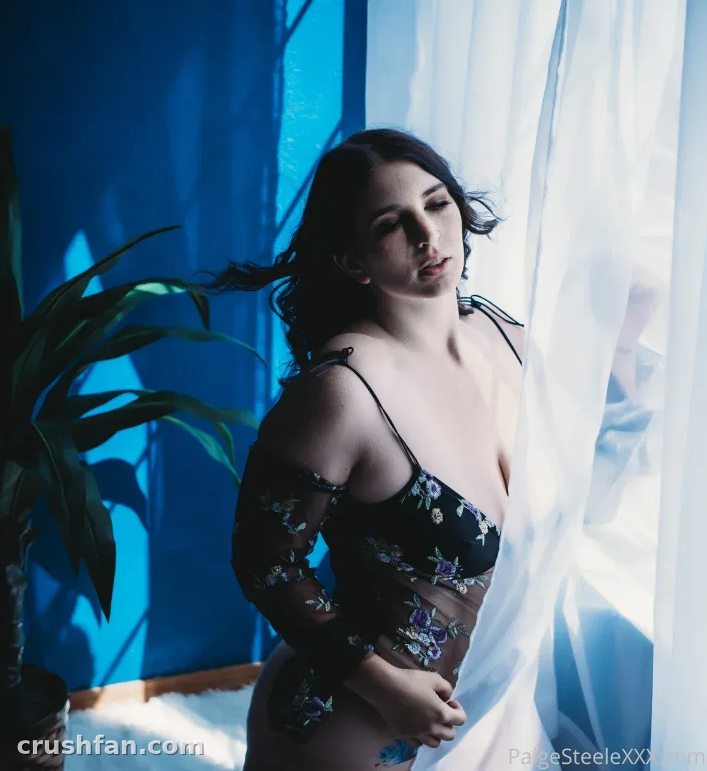 Paige Steele in black floral lingerie beside a bright window with a sheer curtain