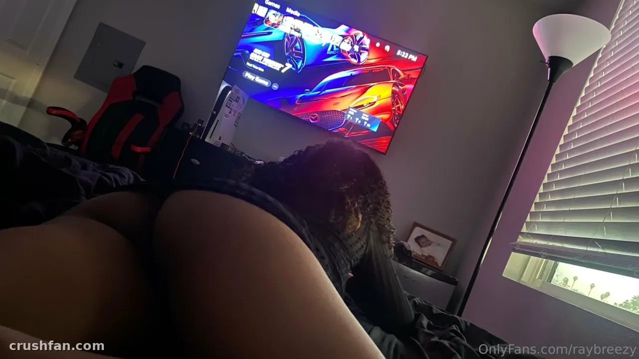 Ray Breezy sentada en una sala de juegos con poca luz, con una pantalla brillante y una lámpara de pie