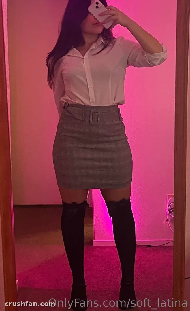 Ingrid Gonzalez prenant un selfie miroir dans une chemise blanche et une jupe grise sous un éclairage rose.