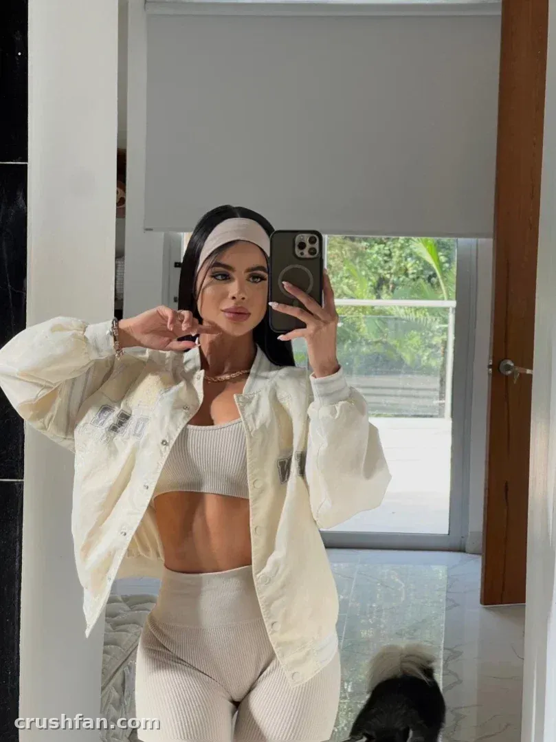 Estefaa tirando uma selfie no espelho com uma jaqueta creme e um conjunto esportivo branco.