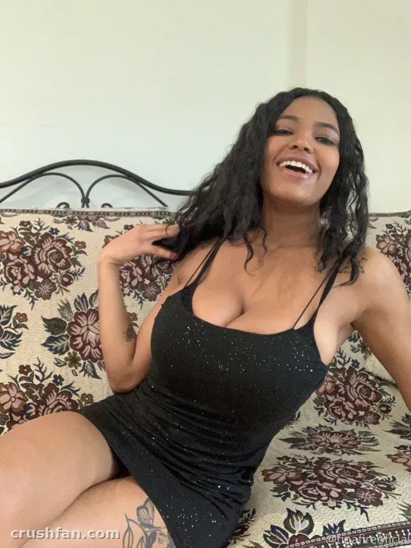 Tina Fire laughing while sitting on a floral couch in a black sparkly dress