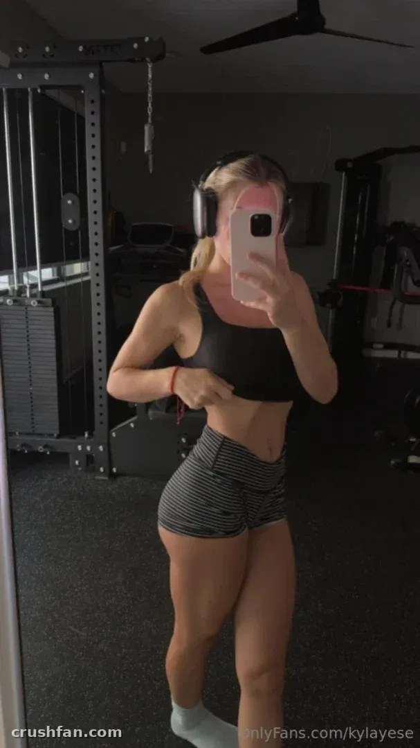 Kyla in einem schwarzen Sport-BH und gestreiften Shorts, die in einem Home-Gym ein Spiegel-Selfie macht.