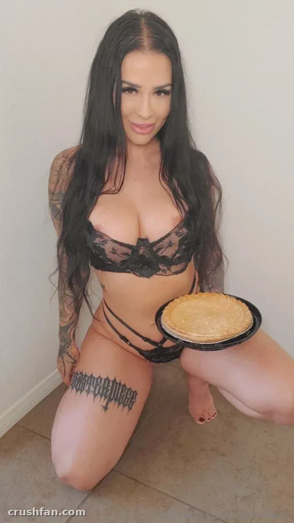 Katrina Jade стоит на коленях в помещении в чёрном кружевном белье, держа пирог
