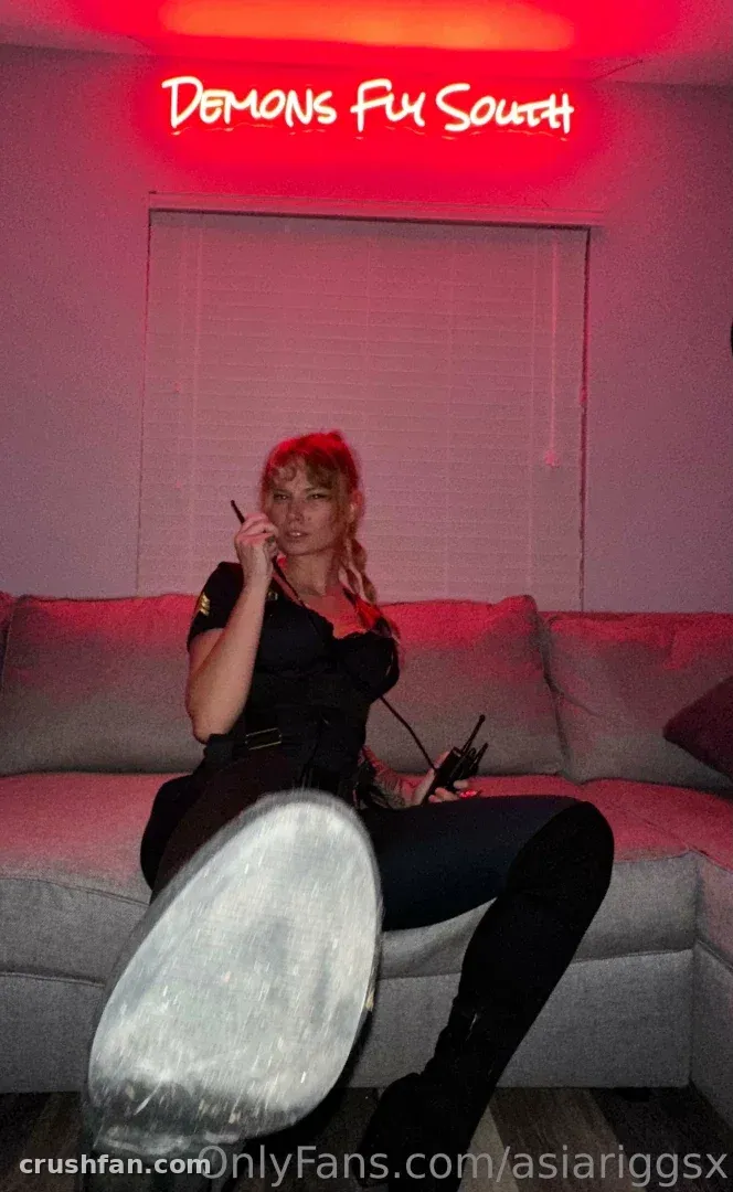 Asia Riggs sentada em um sofá cinza sob um letreiro de neon vermelho, com uma bota levantada em direção à câmera