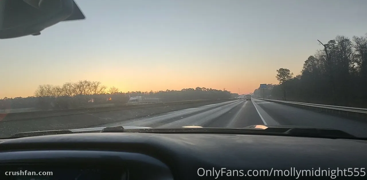 View through a car windshield of a highway at sunrise with bare trees along the shoulder