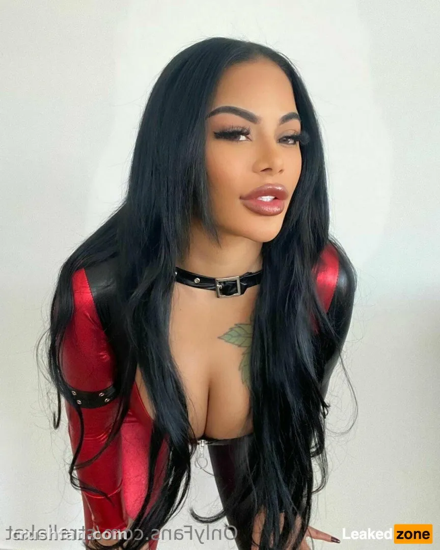Strellakat dans une tenue en latex rouge et noir brillant contre un mur uni